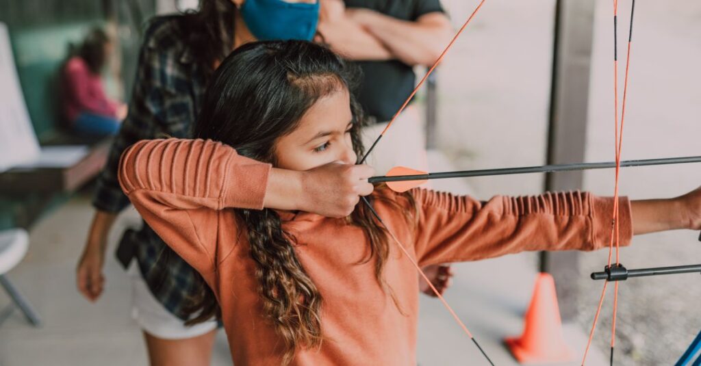 A young girl engaged in archery training outdoors, guided by an adult.